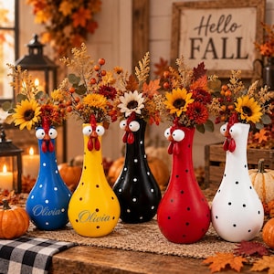 Può includere: Cinque vasi in ceramica a forma di gallina in blu, giallo, nero, rosso e bianco, ciascuno con pois neri e una testa di gallina scolpita con grandi occhi e cresta rossa. Ogni vaso contiene un bouquet di girasoli, bacche e foglie autunnali.