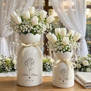 May include: Two white ceramic vases with scalloped rims, each tied with a cream ribbon and filled with white tulips and baby's breath. The larger vase features the text Thalia + Riven and 07. 21. 26, while the smaller displays R + W and 08. 08. 26.