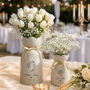 Könnte beinhalten: Zwei weiße Keramikvasen mit Blumengestecken. Eine Vase enthält weiße Tulpen und Schleierkraut, die andere Schleierkraut. Beide Vasen haben einen gewellten Rand, ein Band und ein Blumendesign mit Namen und Daten.