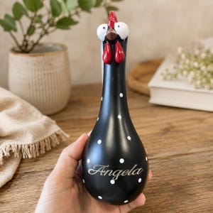 Puede incluir: Una figura de pollo negra en forma de pera con lunares blancos, cresta y barbillas rojas, y ojos blancos grandes. El nombre Angela está grabado en letra cursiva en la parte inferior frontal del cuerpo liso y redondeado del objeto.