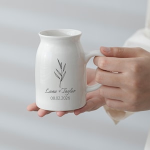 Op de afbeelding: Een witte keramische mok met een afgerond lichaam en een handvat aan de zijkant. De voorzijde toont een zwarte lijntekening van een takje boven de tekst "Luna + Taylor" en de datum "08.02.2026" in een zwart schreeflettertype.