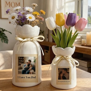 Könnte beinhalten: Zwei weiße Keramikvasen mit gewelltem Rand und cremefarbenen Schleifen. Die größere Vase enthält einen Strauß aus Gänseblümchen und Wildblumen sowie ein Foto eines Vaters mit Kindern und dem Text "I Love You, Dad". Die kleinere Vase enthält vier Tulpen und ein Foto mit dem Text "I'm so grateful for you".