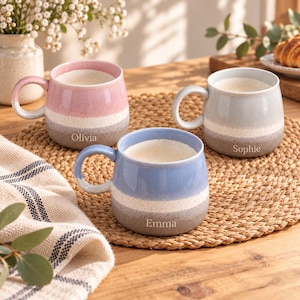 May include: Three ceramic mugs with a speckled finish, each featuring a two-tone colour design. The pink mug is personalised with the name Olivia, the light blue mug with Emma, and the light grey mug with Sophie. Each mug has a rounded handle.
