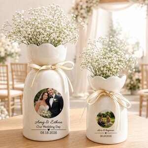 Op de afbeelding: Twee witte keramische vazen met geschulpte randen, elk gestrikt met een crèmekleurig lint en gevuld met gipskruid. De grotere vaas bevat een ronde foto van een koppel en de tekst Amy & Ethan Our Wedding Day 08.18.2026. De kleinere vaas heeft een foto.