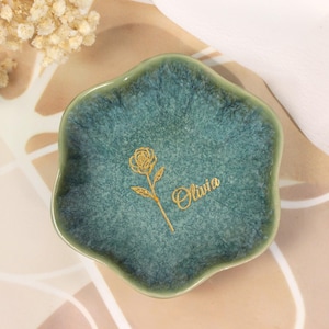 May include: A scalloped, teal-colored ceramic trinket dish with a textured, mottled finish. The center features a gold-colored line art illustration of a single rose stem next to the name Olivia written in a cursive script font.
