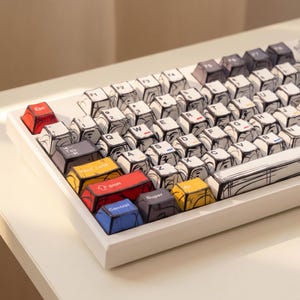 May include: A white mechanical keyboard with a unique comic book style design. The keycaps are white with black outlines, giving them a hand-drawn appearance. Colorful keys include red, yellow, and blue. The keyboard sits on a white surface.