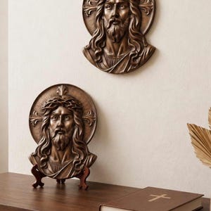 May include: Bronze-toned wall art and bust depicting Jesus with a crown of thorns and a halo. One piece is mounted on the wall, and another rests on a wooden stand. A closed book with a cross is on a wooden shelf.
