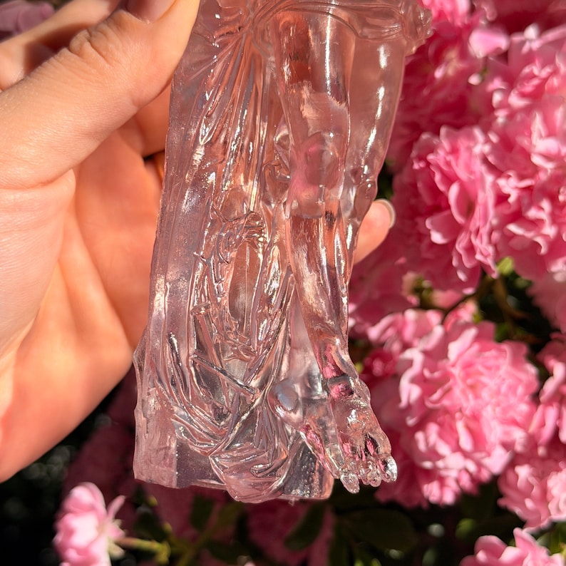 May include: A translucent, pale pink carved crystal figurine depicting a human leg and foot with detailed musculature and draped fabric textures. The sculpture has a smooth, polished surface that catches the light, revealing internal inclusions.