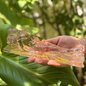 May include: A translucent, light pink glass figurine depicting a reclining figure resting on a rectangular base. The sculpture features detailed contours of a human form lying horizontally, with smooth, reflective surfaces and a clear, pale hue.