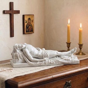 May include: A white marble sculpture of a reclining figure, possibly Jesus, on a rectangular base. A wooden cross and a framed religious icon hang on the wall. Two lit candles in brass holders are on a wooden surface.