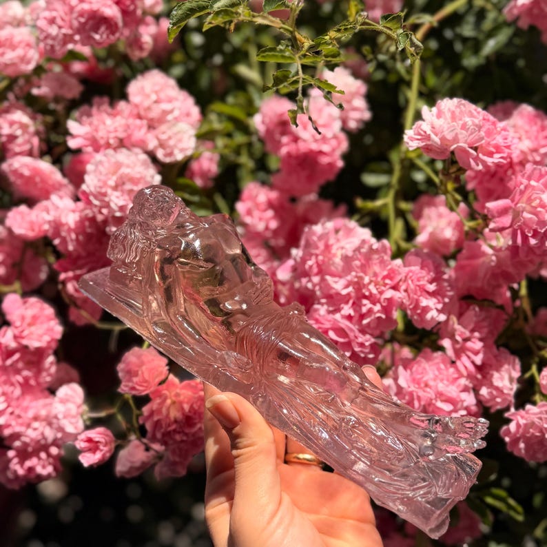 May include: A translucent, light pink glass figurine depicting a reclining figure in a relaxed pose. The sculpture features smooth, molded contours and a clear, glossy finish that catches the light, showcasing the detailed form of the body.