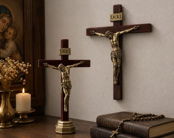 Puede incluir: Dos crucifijos con cruces de madera marrón oscuro y figuras de Jesús en metal color bronce. Cada cruz muestra una pequeña placa rectangular en la parte superior con las letras I.N.R.I. inscritas. Un crucifijo se sostiene sobre una base.