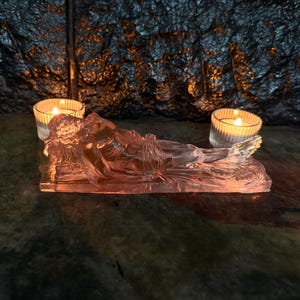 May include: A translucent, pink-tinted glass sculpture depicting a reclining figure resting on a rectangular base. The figure is shown in a horizontal position with detailed contours, illuminated by two small, clear glass votive candle holders.