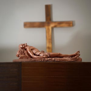 May include: A bronze-colored sculpture of a reclining figure, possibly religious, resting on a rectangular base. A wooden cross is in the background. The sculpture is detailed, with visible facial features and draped fabric.