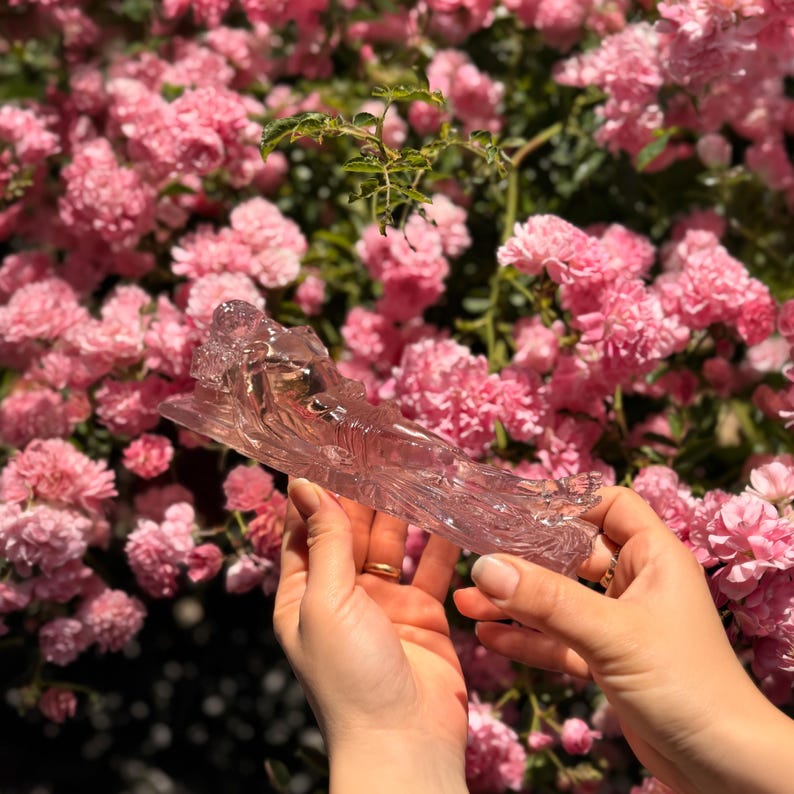May include: A translucent, light pink glass figurine depicting a reclining figure with flowing, textured details. The object is elongated and sculpted with smooth, reflective surfaces that catch the light, showcasing a clear, crystalline appearance.