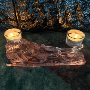 May include: A translucent pink glass sculpture depicting a reclining figure resting on a rectangular base. The figure is positioned horizontally with legs extended. Two clear, ribbed glass votive candle holders with lit white candles flank the sculpture.
