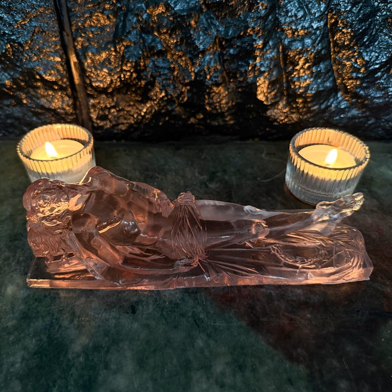 May include: A translucent pink glass sculpture depicting a reclining figure resting on a rectangular base. The figure is positioned horizontally with legs extended. Two clear, ribbed glass votive candle holders with lit white candles flank the sculpture.