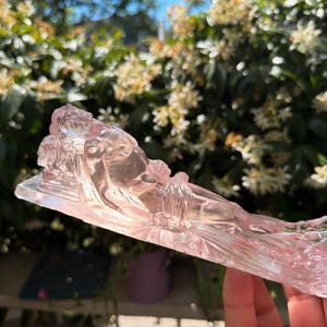 May include: A translucent, light pink glass figurine depicting a reclining figure resting on a rectangular base. The sculpture features smooth, molded contours and a clear, glossy finish that catches the light, showcasing the detailed form of the figure.