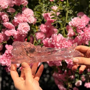 May include: A translucent, light pink glass figurine depicting a reclining figure in a horizontal position. The sculpture features smooth, molded contours and a clear, crystalline appearance, showcasing a detailed form resting on a flat base.