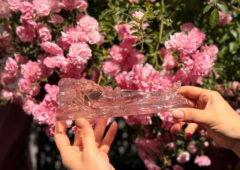 May include: A translucent, light pink glass figurine depicting a reclining figure in a horizontal position. The sculpture features smooth, molded contours and a clear, crystalline appearance, showcasing a detailed form resting on a flat base.