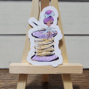 May include: A purple and gold thread spool with a fairy sitting on top. The fairy has purple hair, a blue dress, and a white bow. The fairy is holding a pair of scissors.