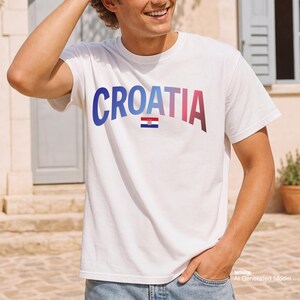 May include: White t-shirt with the word "CROATIA" in blue, pink, and red gradient lettering. The Croatian flag is below the word. The shirt is worn by a person.