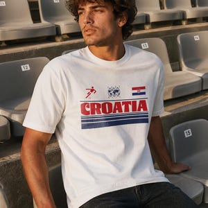 K&ouml;nnte beinhalten: Ein wei&szlig;es kurz&auml;rmeliges T-Shirt mit einem Grafikdruck, der ein rotes Fu&szlig;ballspieler-Symbol, einen Globus, die kroatische Flagge und das Wort CROATIA in fetten roten Buchstaben &uuml;ber zwei horizontalen dunkelblauen Streifen zeigt.