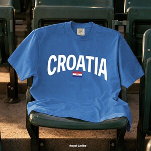May include: A blue t-shirt with the word "CROATIA" in white arched lettering and a small Croatian flag. The shirt is displayed on a green stadium seat. The text "Royal Caribe" is at the bottom.