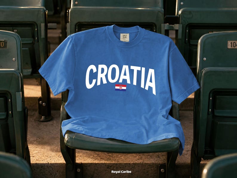 May include: A blue t-shirt with the word "CROATIA" in white arched lettering and a small Croatian flag. The shirt is displayed on a green stadium seat. The text "Royal Caribe" is at the bottom.