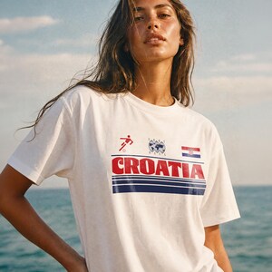 K&ouml;nnte beinhalten: Ein wei&szlig;es kurz&auml;rmeliges T-Shirt mit einem Grafikdruck auf der Brust. Das Design zeigt ein kleines rotes Fu&szlig;ballspieler-Symbol, eine Weltkugel mit dem Text FOOT BALL, die kroatische Flagge und das Wort CROATIA in gro&szlig;en roten Buchstaben &uuml;ber blauen Linien.