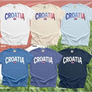 May include: Six short-sleeved t-shirts in various colors, including white, ivory, and blue. Each shirt displays the word "CROATIA" in a curved font, with the Croatian flag beneath the text. The shirts are on a red track and green grass background.