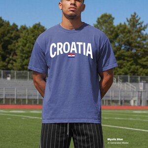 May include: A mystic blue t-shirt with the word "CROATIA" in white block letters across the chest, above a small Croatian flag. The shirt is short-sleeved and worn by a person standing on a green field.