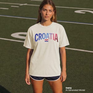 May include: Ivory-colored t-shirt with the word "CROATIA" in a gradient of blue to red. Below the word is a small Croatian flag. The model is wearing navy blue shorts with white trim. The background is a green field.