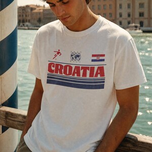 K&ouml;nnte beinhalten: Ein wei&szlig;es kurz&auml;rmeliges T-Shirt mit einem Grafikdruck, der das Wort CROATIA in gro&szlig;en roten Buchstaben, ein kleines rotes Fu&szlig;ballspieler-Symbol, eine Weltkugel-Illustration, den Text FOOT BALL, die kroatische Flagge und drei blaue Streifen zeigt.