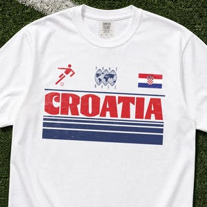 K&ouml;nnte beinhalten: Ein wei&szlig;es Kurzarm-T-Shirt mit einem Grafikdruck, der das Wort CROATIA in gro&szlig;en roten Buchstaben, ein rotes Fu&szlig;ballspieler-Symbol, eine Weltkugel mit dem Text FOOT BALL und die kroatische Flagge &uuml;ber drei horizontalen dunkelblauen Streifen zeigt.