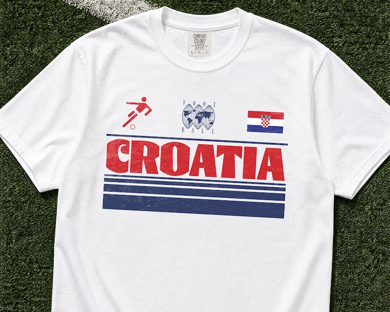 K&ouml;nnte beinhalten: Ein wei&szlig;es Kurzarm-T-Shirt mit einem Grafikdruck, der das Wort CROATIA in gro&szlig;en roten Buchstaben, ein rotes Fu&szlig;ballspieler-Symbol, eine Weltkugel mit dem Text FOOT BALL und die kroatische Flagge &uuml;ber drei horizontalen dunkelblauen Streifen zeigt.