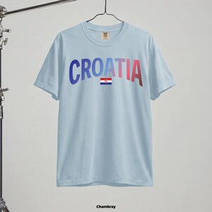 May include: Light blue t-shirt with the word "CROATIA" in gradient blue to red lettering and a small Croatian flag. The shirt is hanging on a hanger. The word "Chambray" is at the bottom.