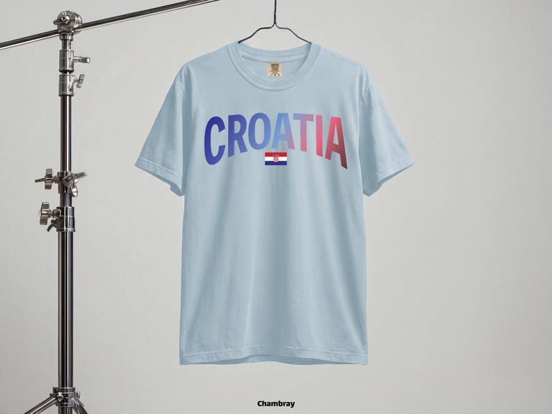 May include: Light blue t-shirt with the word "CROATIA" in gradient blue to red lettering and a small Croatian flag. The shirt is hanging on a hanger. The word "Chambray" is at the bottom.
