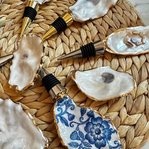 May include: A collection of bottle stoppers featuring natural oyster shells with iridescent white interiors and gold-painted edges. One stopper displays a blue and white floral pattern on the shell surface. Each includes a metal base with black rings.