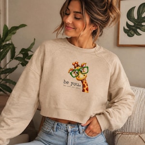 Può includere: Una felpa corta color beige a maniche lunghe con la grafica di una giraffa che indossa occhiali verde brillante. Sotto la giraffa, il testo recita "be you." seguito dal testo più piccolo "The world will adjust." La felpa ha un girocollo.