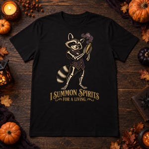 May include: A black t-shirt featuring a graphic of a raccoon dressed in a skeleton costume and a purple waistcoat, holding a cocktail shaker with purple smoke rising from it. Below the raccoon, the text reads, I SUMMON SPIRITS FOR A LIVING.