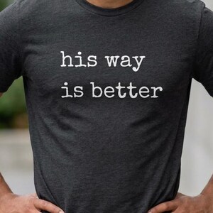 May include: Dark grey t-shirt with the white text "his way is better". The t-shirt is a crew neck style and appears to be made of a soft material. The person is standing outside in front of a blurred background.