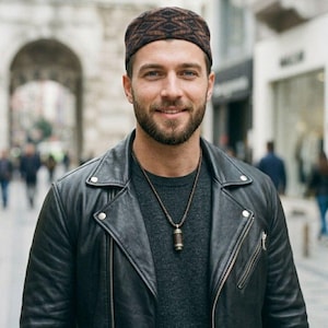 May include: A man in a black leather jacket, grey sweater, and a brown patterned cap. He is also wearing a pendant necklace. The background shows a blurred street scene with buildings and people.