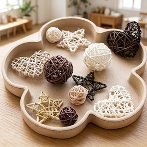 Può includere: Un vassoio beige a forma di nuvola contenente un assortimento di oggetti decorativi in rattan intrecciato, tra cui sfere, stelle e cuori bianchi, marroni e beige disposti sulla superficie interna del vassoio.