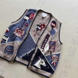 Peut inclure: Un gilet en tricot sans manches présentant un motif patchwork dans des tons beige, bleu et marine. Le vêtement affiche divers motifs brodés, notamment une tête de cheval blanche, des ballons et des formes géométriques, avec une fermeture boutonnée.