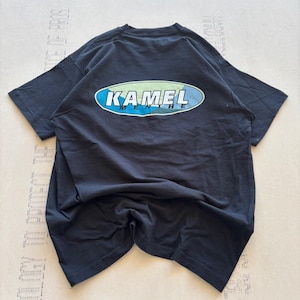 May include: A black short-sleeve t-shirt featuring a central graphic on the back. The graphic consists of a horizontal oval shape with a light green and blue background, containing the white text KAMEL MENTHE in a bold, sans-serif font.