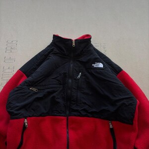 May include: A red and black zip-up jacket featuring a high collar, long sleeves, and a fleece texture. The upper chest and shoulder panels are black, while the lower body and sleeves are red. It includes a white The North Face logo on the chest.
