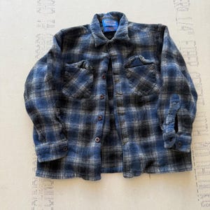 Pode incluir: Uma camisa de manga comprida com botões e um padrão xadrez em azul, cinzento e preto. A camisa inclui um colarinho pontiagudo, dois bolsos no peito com abas e botões escuros na frente. Uma etiqueta azul da marca Pendleton é visível no pescoço.
