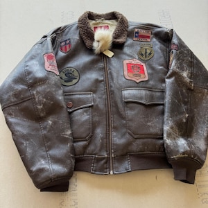 Puede incluir: Una chaqueta bomber de cuero marrón con cuello de piel sintética marrón, cierre de cremallera frontal y puños acanalados. La chaqueta está decorada con múltiples parches bordados, incluyendo una bandera de Texas, un emblema Cobra y la insignia de la U.S. Air Force.