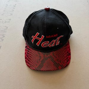Pode incluir: Um boné de beisebol preto com o texto Miami Heat bordado em vermelho e branco na parte frontal. O boné possui uma aba com padrão de pele de cobra em vermelho e preto e um pequeno botão vermelho no topo central da coroa.